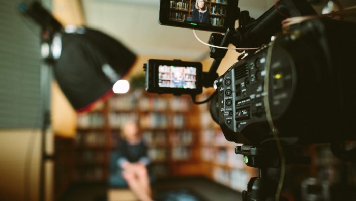 A video camera in the foreground recording a woman sitting in front of bookcases in the background.