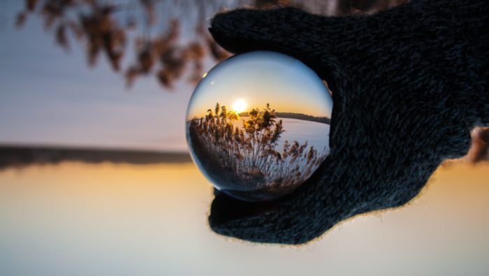 A gloved hand in the foreground holding a crystal ball which shows an inverted image of the scenic background of a sunrise.