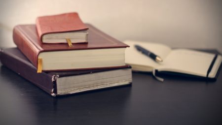 Photograph of a stack of three leather-bound notebooks alongside an open notebook with a pen on a dark table.