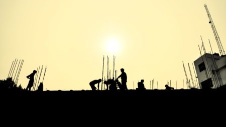 A photo showing silhouettes of people doing construction work against a pale yellow sky.