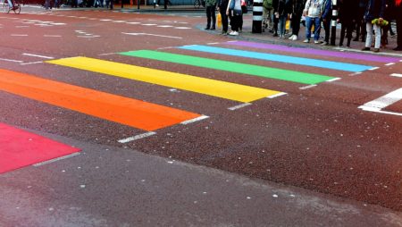The photo shows a cross walk in pride colours in an urban setting. People are waiting to cross the street.