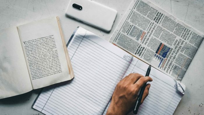 The picture displays a hand that is writing into an open notebook. A pjone and news paper are next to it.