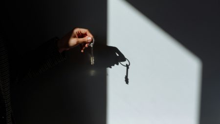 A hand holding a set of keys casts a sharp shadow on a wall illuminated by a bright, angled beam of light, creating a high-contrast scene.