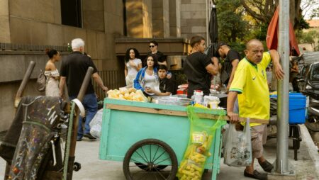 A street scene with a vendor leaning against a pole beside a turquoise cart filled with corn on the cob, while a small crowd gathers around on a sidewalk near a building.